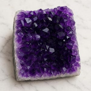 May include: A rectangular amethyst geode with a crystalline structure. The crystals range in color from deep purple to light lavender. The geode is set against a white marble surface.