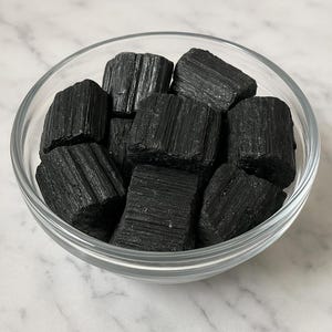 Puede incluir: Un cuenco de vidrio transparente lleno de cristales de turmalina negra en bruto. Los cristales tienen un aspecto texturizado y estriado. El cuenco se encuentra sobre una superficie de m&aacute;rmol blanco y gris.