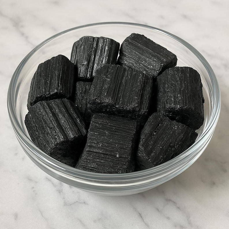 Puede incluir: Un cuenco de vidrio transparente lleno de cristales de turmalina negra en bruto. Los cristales tienen un aspecto texturizado y estriado. El cuenco se encuentra sobre una superficie de m&aacute;rmol blanco y gris.