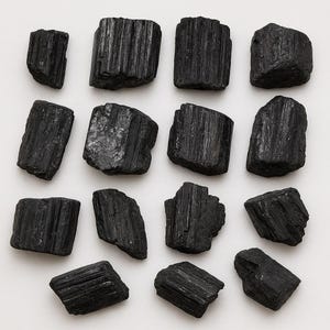 Puede incluir: Una colecci&oacute;n de cristales de turmalina negra en bruto. Los cristales tienen una superficie rugosa y texturizada con estr&iacute;as verticales. Las piedras var&iacute;an en tama&ntilde;o y forma, dispuestas sobre un fondo blanco. Estas piedras preciosas naturales se utilizan a menudo por sus propiedades metaf&iacute;sicas.