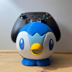 May include: A blue, white, and yellow Piplup-shaped controller holder with a black controller resting on top. The holder is on a wooden surface, with a light grey background.