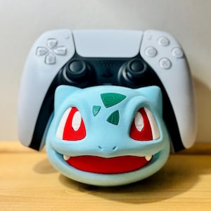 Puede incluir: Un soporte para mando azul claro con diseño de Bulbasaur, con ojos y boca rojos, y detalles verdes. Un mando de PlayStation 5 blanco y negro se coloca detrás de la figura de Bulbasaur. Para entusiastas de los videojuegos.