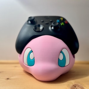 May include: A pink, cartoon character with large blue eyes and a black Xbox controller on top. The character has white accents around the eyes and a rounded shape, sitting on a wooden surface.