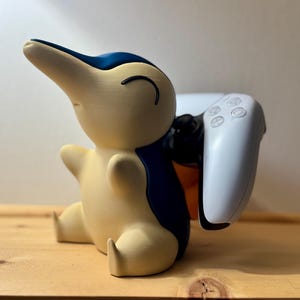 May include: A cream-coloured, cartoon-style figure with blue accents, holding a white game controller. The figure is resting on a wooden surface. The controller is white with black buttons.