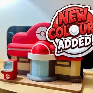 May include: A red and tan model of a building with a sign that says "NEW COLOURS ADDED!". The model has a red roof and a blue and white circular design. There is a small red arcade machine on the left side of the model.