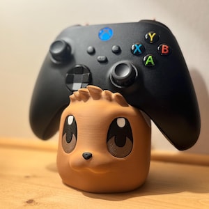 May include: A black Xbox controller rests on a brown, cartoon-style character holder. The controller has colored buttons and a blue Xbox logo. The holder has large eyes and a small nose.
