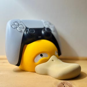 Op de afbeelding: Een gele Psyduck-figuur met een lichtgrijze en zwarte PlayStation 5-controller die op zijn hoofd rust. De eend heeft een beige snavel en witte ogen. De achtergrond is een lichtbruin houten oppervlak en een lichtbeige muur.