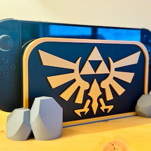Könnte beinhalten: Ein schwarzer und goldener dekorativer Ständer für eine Spielkonsole mit einem stilisierten Emblem. Der Ständer wird von zwei grauen geometrischen Formen getragen. Eine schwarze Spielkonsole befindet sich im Ständer.