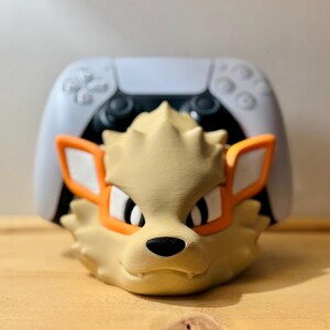 Può includere: Una testa di personaggio stilizzata color beige con occhiali con montatura arancione, occhi neri e naso nero. Il personaggio è davanti a un controller di gioco bianco e nero. Il personaggio ha una criniera a spillo.