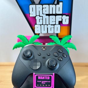 May include: A black video game controller resting on a stand featuring the Grand Theft Auto VI logo in blue, pink, and orange. The stand includes two green palm trees and a pink sign with the text WANTED, YOUR GAMERTAG HERE, and five stars.