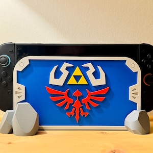 Könnte beinhalten: Eine schwarze Nintendo Switch-Konsole mit einem blauen, weißen, gelben und roten dekorativen Schild mit einem stilisierten Emblem. Das Schild wird von zwei grauen, felsförmigen Ständern getragen.