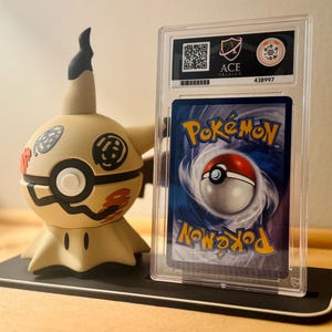 May include: A Mimikyu figure stands beside a graded Pokémon card in a protective case. The Mimikyu figure is tan with black and red accents. The Pokémon card features a Poké Ball design and the word "Pokémon". The case has a label with the text "ACE Gaming".
