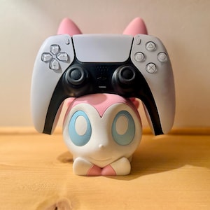 May include: A white and black PlayStation 5 controller rests on a pink and white Sylveon-themed controller stand. The stand has blue eyes and pink ears. The controller has a black center and white buttons.