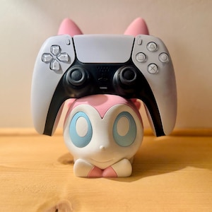 May include: A white and black PlayStation 5 controller rests on a pink and white character-themed stand. The stand has a cartoonish design with blue eyes and pink accents, and is sitting on a wooden surface.