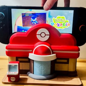 Pode incluir: Um suporte vermelho e branco impresso em 3D do Centro Pokémon segurando um console Nintendo Switch preto exibindo o jogo "Pokémon Pokepia". O suporte tem um design detalhado com uma pequena máquina de arcade vermelha.