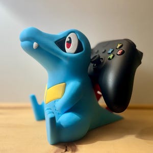 Può includere: Un supporto blu a forma di Totodile per controller con un controller Xbox nero. Il supporto ha una toppa gialla sul lato e dettagli bianchi. Il controller è nero con pulsanti colorati.