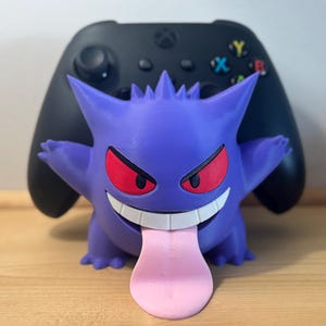 May include: A purple Gengar-shaped controller holder with red eyes and a pink tongue, in front of a black Xbox controller. The holder has a wide grin and spiky details. The background is a wooden surface.