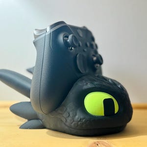 Könnte beinhalten: Schwarzer Controller-Ständer in Form eines Drachenkopfes mit einem neongrünen Auge. Ein Xbox-Controller ruht in dem Ständer. Der Ständer hat eine strukturierte Oberfläche und ist so konzipiert, dass er den Controller aufrecht hält.