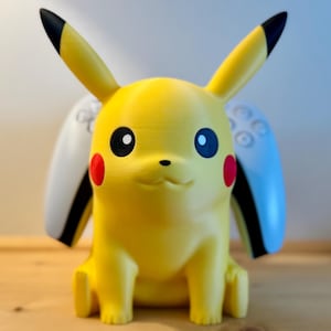 May include: A yellow Pikachu figurine with black-tipped ears, red cheeks, and black eyes. A white and blue game controller is behind the figure. The figure is sitting on a wooden surface.