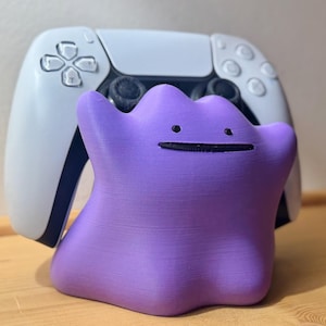 May include: A purple Ditto figurine with a simple black facial expression, positioned in front of a white and black game controller. The controller has a symmetrical design with black buttons and a central touchpad. The background is a light wooden surface.