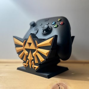 Peut inclure: Manette de jeu noire posée sur un support noir et doré. Le support présente un motif doré. La manette a des boutons colorés. Le support est sur une surface en bois.