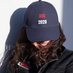 Op de afbeelding: Marineblauwe baseballcap met de tekst "AOC 2028" in rood en wit. De cap is een klassieke stijl, geschikt voor casual wear. De afbeelding toont de cap op een persoon.