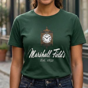 May include: A woman wearing a dark green t-shirt with the text "Marshall Field's Est. 1852" in white script. The shirt features a graphic of a clock inside a wooden frame. She is wearing dark blue jeans.
