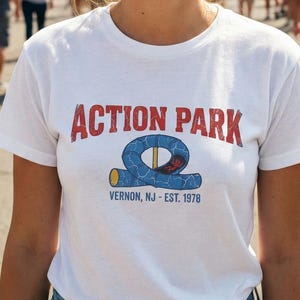 May include: A white t-shirt with the words "ACTION PARK" in red block letters and a blue and yellow graphic. Below the graphic is the text "VERNON, NJ - EST. 1978". The shirt is worn by a person in a crowd at an amusement park.