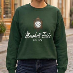 May include: A dark green sweatshirt featuring a clock design and the text "Marshall Field's Est. 1852". The sweatshirt is a crew neck style and is made of a soft material. The design is in white and brown.