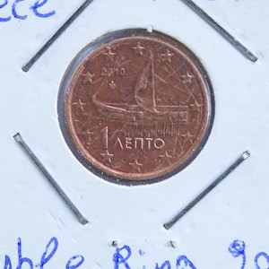May include: A copper 1 Euro cent coin from Greece, dated 2010, depicting a sailing ship. The coin is held in a protective sleeve with handwritten text: "Greece" and "Double Ring 2010".