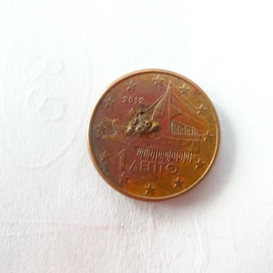 Może przedstawiać: Miedziana moneta 1 eurocent z 2012 roku. Moneta przedstawia wzór żaglowca z napisem "1 ΛΕΠΤΟ" i jest otoczona gwiazdami. Moneta znajduje się na białym tle.