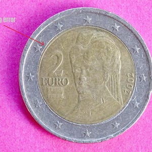 Puede incluir: Una moneda de dos euros con un centro dorado y un anillo exterior plateado, que presenta un retrato y el número "2" con la palabra "EURO". La moneda muestra un error de acuñación, con el texto "Minting error" que señala el defecto.