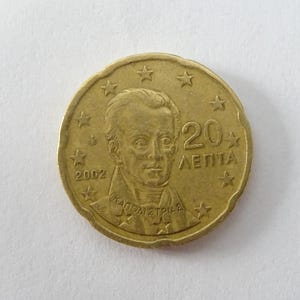 May include: A gold-colored coin featuring a portrait of a man, the year 2002, and the text "20 ΛΕΠΤΑ". The coin is surrounded by stars and has a slightly worn edge. The coin is a collectible item.