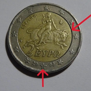 Op de afbeelding: Een munt van twee euro met een gouden kern en een zilveren buitenring. De gouden kern toont een vrouw die een stier berijdt, met de tekst "2 EYPΩ". De rand van de munt heeft sterren en het jaar "2002".