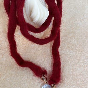 May include: A handcrafted necklace with a deep red, felted wool cord. The cord is looped and features two large, irregular, white baroque pearls. The pearls are attached with gold-coloured wire and small gold beads. The necklace is displayed on a neutral-toned surface.