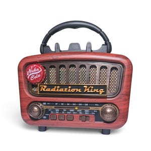 May include: A portable radio with a faux wood grain finish, featuring a black top handle and a speaker grille. The front displays a Nuka Cola bottle cap emblem, a Radiation King logo, two circular control knobs, and a frequency tuning scale.