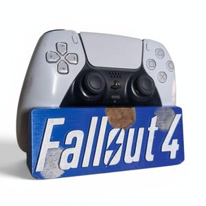 Op de afbeelding: Een witte en zwarte PlayStation 5-controller rust op een blauwe houten basis. De basis toont de witte tekst "Fallout 4" met een blikseminslag. De controller heeft een zwart middengedeelte en witte zijkanten.
