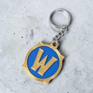 Könnte beinhalten: Ein blau-goldener Schlüsselanhänger mit einem stilisierten Buchstaben "W" in einem kreisförmigen Emblem. Er hat eine silberne Kette und einen Ring. Das Design hat einen leuchtend blauen Hintergrund mit goldenen Details.