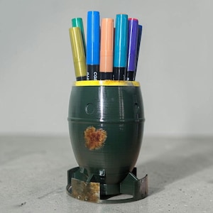 Może przedstawiać: Ciemnozielony pojemnik na długopisy w kształcie bomby, z żółtą obwódką i rdzawym wzorem w kształcie serca. Zawiera kilka kolorowych markerów, w tym złoty, niebieski i pomarańczowy. Pojemnik na długopisy stoi na szarej powierzchni.