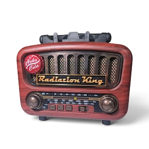 May include: A tabletop radio with a faux wood grain finish, featuring a black speaker grille and a gold-colored plaque reading Radiation King. A red Nuka-Cola bottle cap is attached to the front. The radio includes two circular knobs and a frequency display panel with AM, FM, and SW bands.