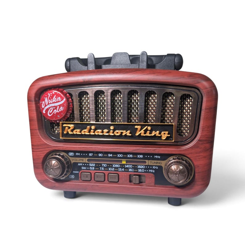 May include: A tabletop radio with a faux wood grain finish, featuring a black speaker grille and a gold-colored plaque reading Radiation King. A red Nuka-Cola bottle cap is attached to the front. The radio includes two circular knobs and a frequency display panel with AM, FM, and SW bands.
