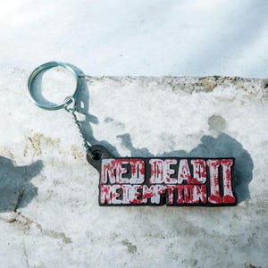 Könnte beinhalten: Ein Schlüsselanhänger mit dem Text "RED DEAD REDEMPTION II" in roter und weißer Schrift auf schwarzem Hintergrund. Er hat einen silbernen Schlüsselring und eine Kette.