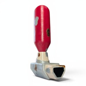 Puede incluir: Un inhalador rojo y plateado con una parte superior cilíndrica roja y una base plateada. El inhalador tiene un aspecto desgastado y envejecido con pintura descascarillada y manchas de óxido. La base tiene forma rectangular con una abertura negra.