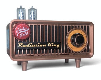 May include: A copper-colored radio with a black speaker grille, featuring the text Radiation King in gold lettering. A red Nuka Cola bottle cap is attached to the front. Two clear glass vacuum tubes sit on top, and a circular dial is on the right.