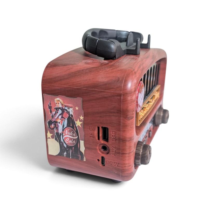 May include: A portable speaker with a faux wood grain finish, featuring a sticker of a character next to a Nuka Cola bottle. The device includes a black top handle, a front speaker grille, two control knobs, and side ports for TF, USB, AUX, and DC 5V.