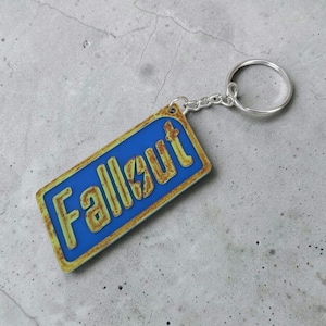 Könnte beinhalten: Rechteckiger Schlüsselanhänger mit dem Wort "Fallout" in Gelb auf blauem Grund. Er hat einen silbernen Schlüsselring und eine Kette. Die Ränder haben ein verwittertes, rostiges Aussehen. Ein Gaming-Accessoire.