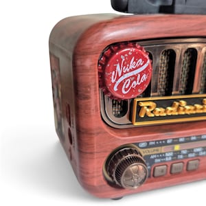 May include: A portable radio with a faux wood grain finish, featuring a red Nuka Cola bottle cap attached to the front speaker grille. The device includes a gold-colored Radiance label, a circular volume knob, and a frequency display panel.