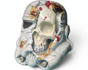 May include: A white and gray Stormtrooper helmet with a skull design. The helmet is weathered with brown and red accents, suggesting damage. The skull is visible through the damaged helmet, with visible teeth and eye sockets. The overall theme is a blend of science fiction and macabre.