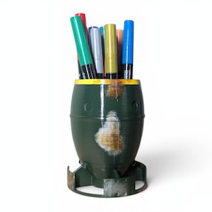 Pode incluir: Um porta-canetas verde em forma de bomba, com uma borda amarela e um aspeto enferrujado. Contém vários marcadores coloridos, incluindo verde, vermelho, azul e dourado. Mede cerca de 15 cm de altura.