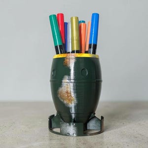 May include: A dark green pen holder shaped like a bomb, with a yellow rim and a rusty appearance. The pen holder contains several colorful pens, including green, red, blue, and orange. The base of the holder has a fin-like design.